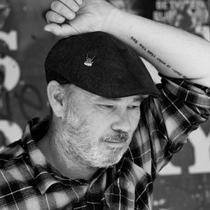 A black-and-white portrait headshot of a man wearing a hat with his arm raised above his head.
