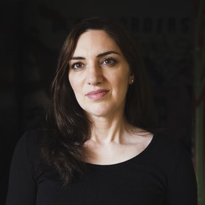 A portrait headshot of a woman with dark hair and a black boatneck top against a black background.