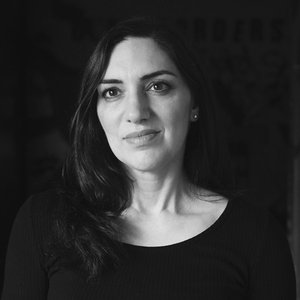 A portrait headshot of a woman with dark hair and a black boatneck top against a black background.