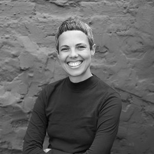 A portrait headshot of a poet in a high-neck sweater and cropped hair against a textured wall, smiling.