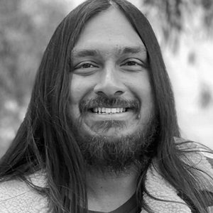 A black and white portrait headshot of a man with long hair and a beard, smiling.