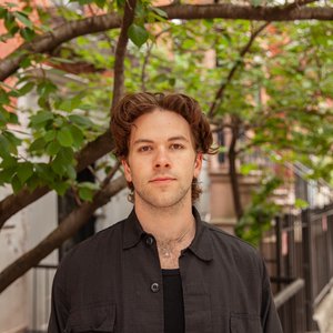 A headshot showing the subject in a dark coat standing on a residential street with greenery and buildings in the background.