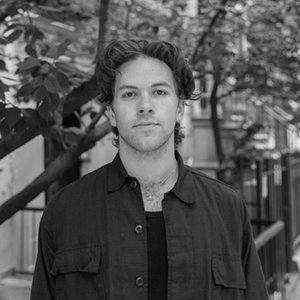 A black-and-white headshot where the poet is standing in a tree-lined street, looking into the camera.