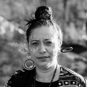 A portrait of a First Nations woman with woven earrings and ceremonial face paint, standing outdoors.