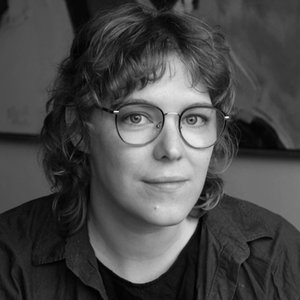 A portrait headshot of a poet with shoulder-length hair and glasses, wearing a collared shirt.