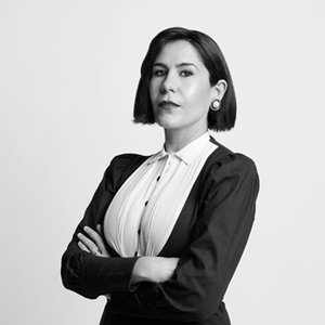A black-and-white headshot of a woman in a blazer with her arms folded against a white background.