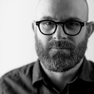A black-and-white headshot of a man with a beard, glasses, and dark collared shirt against a white  background.
