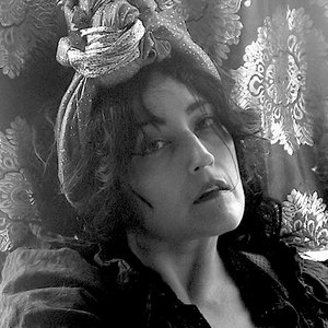 A black-and-white headshot of an artist against a patterned tapestry.