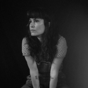 A black-and-white headshot of a woman wearing a striped shirt and overalls, looking at something to the left of the frame.