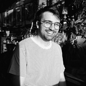 A portrait headshot of a poet in a striped shirt sitting in the sun.