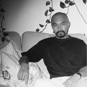 A black-and-white headshot of a man sitting on a couch wearing a dark shirt with the leafy vine of a plant behind him.