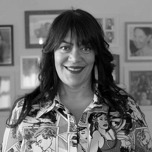 A woman standing in front of a wall of framed artwork, smiling, wearing a shirt with an old-school comic strip print.