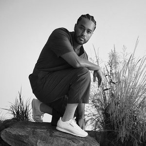 A black-and-white shot of a man crouching on a rock among the grasses, looking down into the camera.