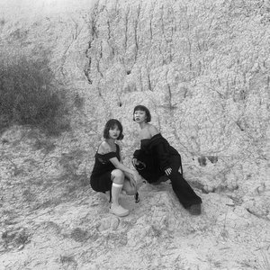A black-and-white shot of two people squatting in what appears to be dry, cracked earth or clay. They are in the middle of the frame, and dry grass is to the top left.